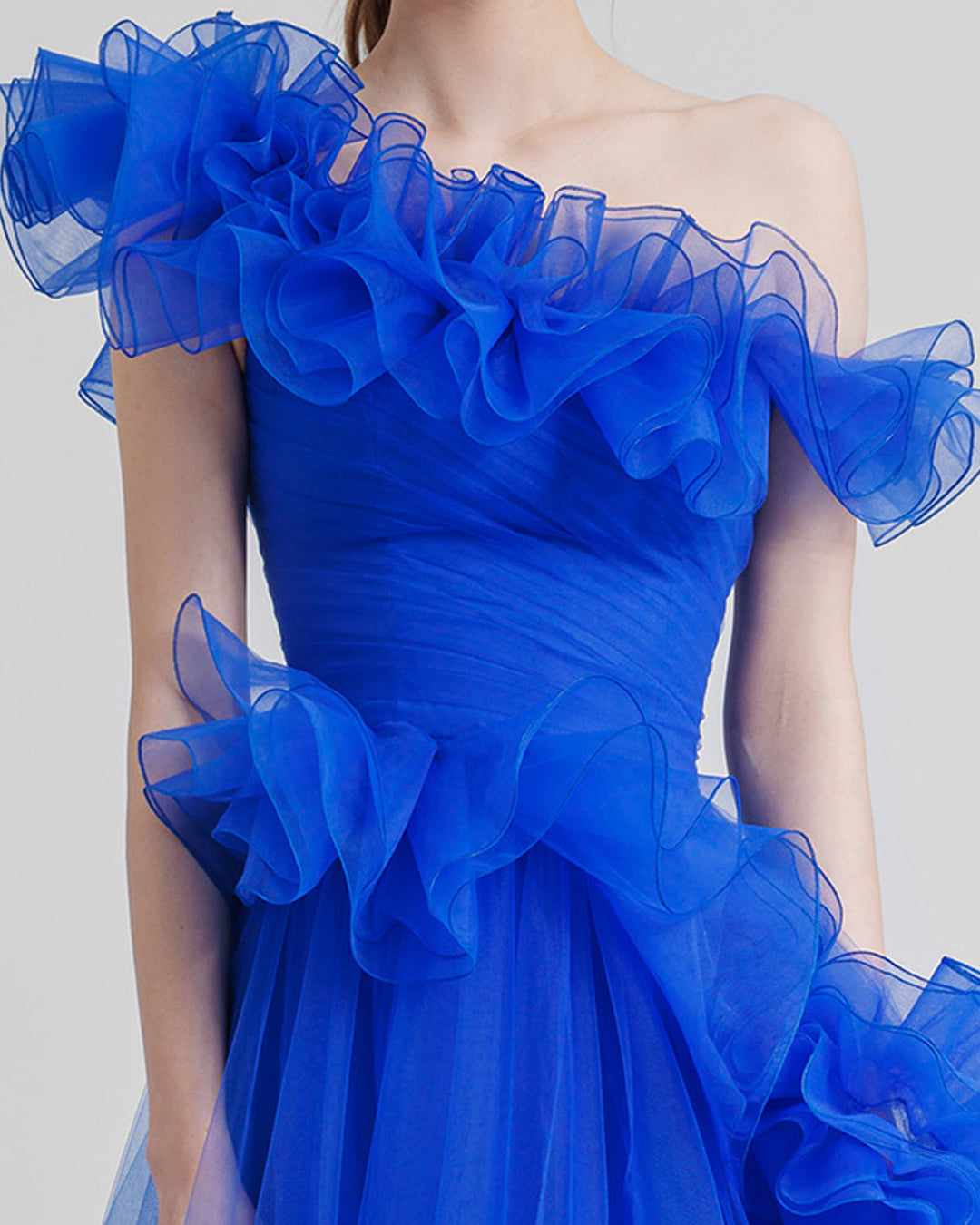 A close-up of a flared royal blue evening dress featuring an asymmetrical ruffled neckline, and ruffled embellishments on the skirt.