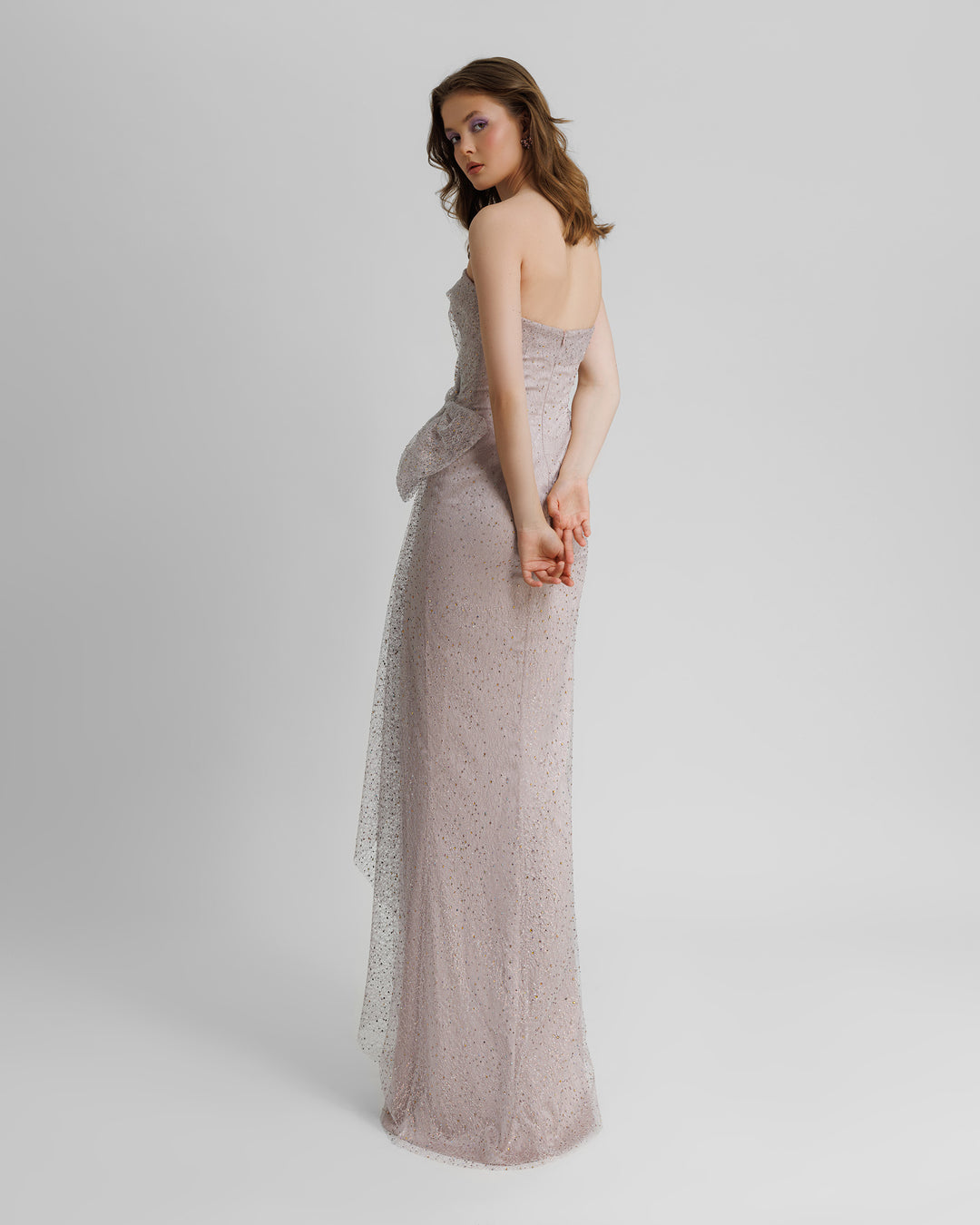 The back of a strapless dubetti-like bodice long lilac evening dress with draping details, a bow design on the waist, and a slit on the side.