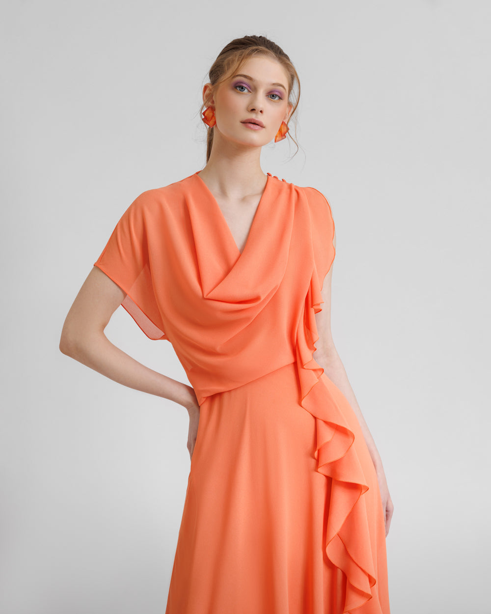 A close-up of an asymmetrical flared orange dress with draping and ruffled details on the side.