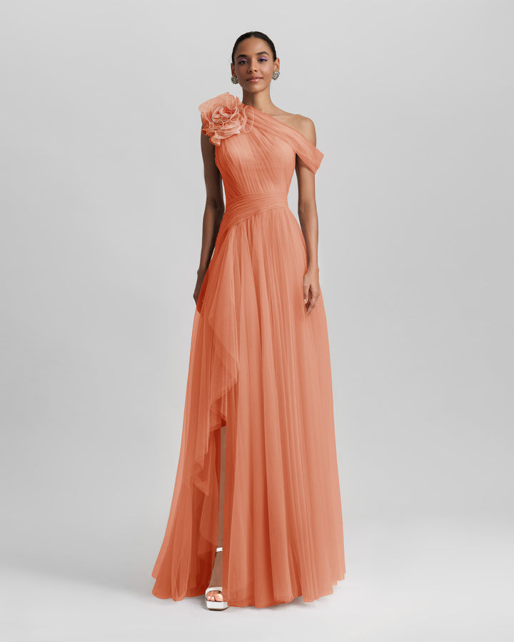 An asymmetrical neckline full tulle long evening dress in peach color, with a flower design on the shoulder, a ruffled side slit, and a draping design on the bodice.