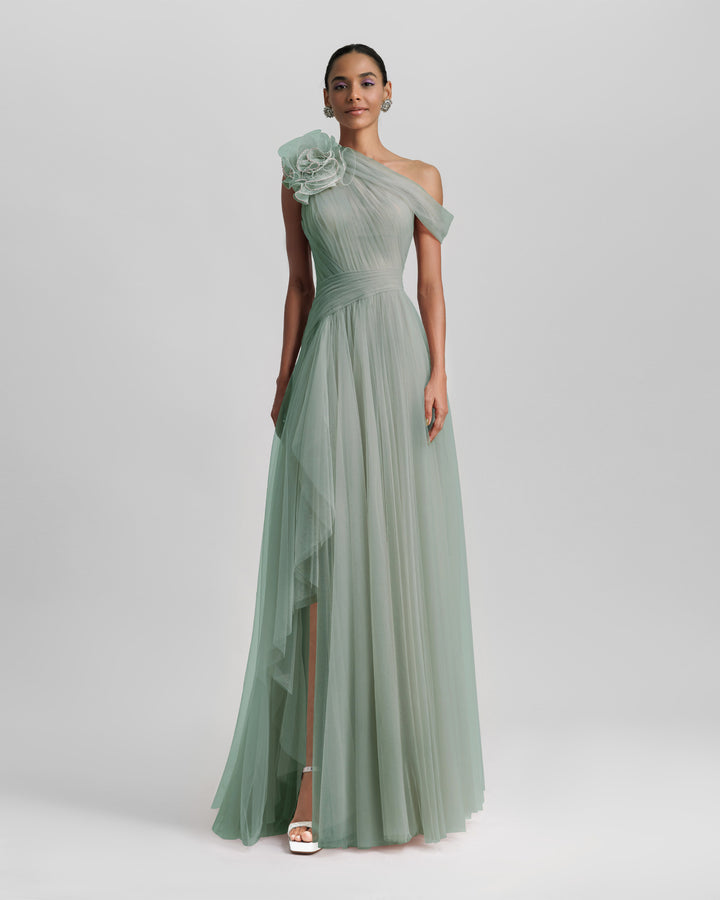 A full tulle long evening dress in mint color, with an asymmetrical neckline, a flower design on the shoulder, a ruffled side slit and a draping design on the bodice.