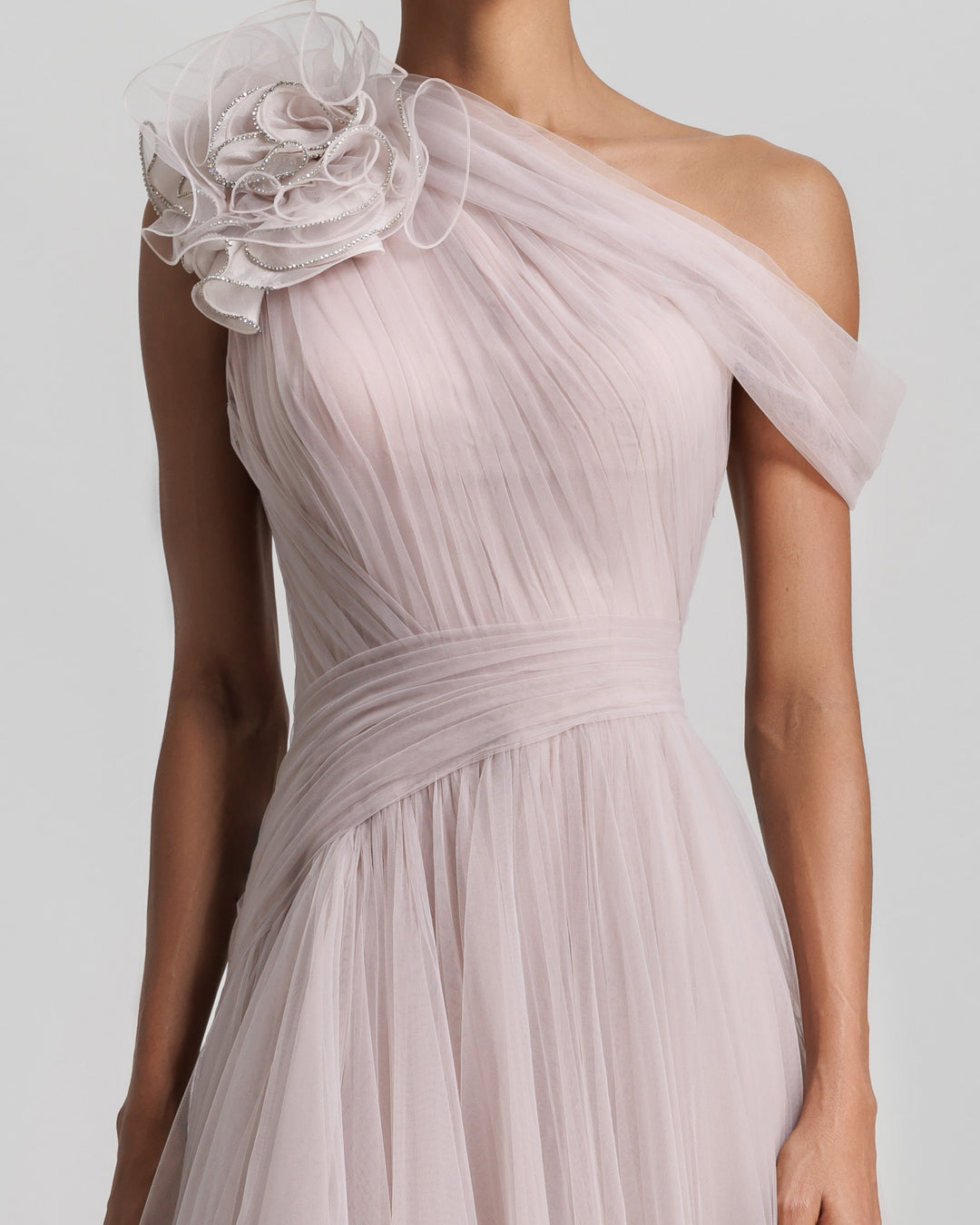 A close-up of a full tulle long evening dress, with an asymmetrical neckline, a flower design on the shoulder, a ruffled side slit, and draping design on the bodice.