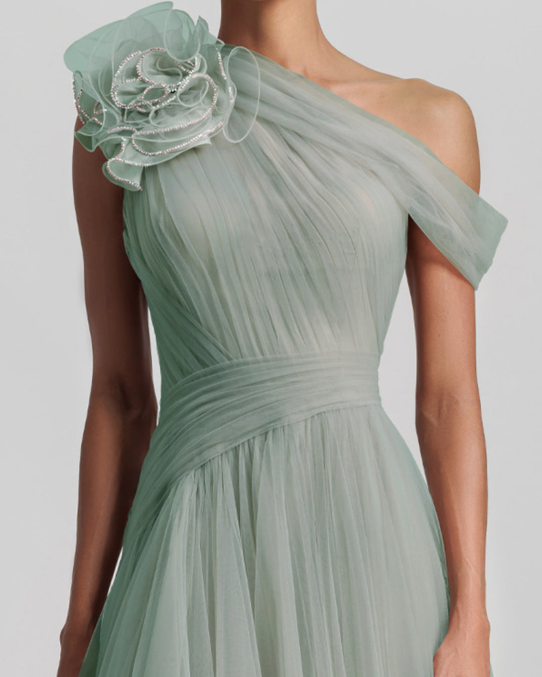 A close-up of a full tulle long evening dress in mint color, with an asymmetrical neckline, a flower design on the shoulder, a ruffled side slit and a draping design on the bodice.