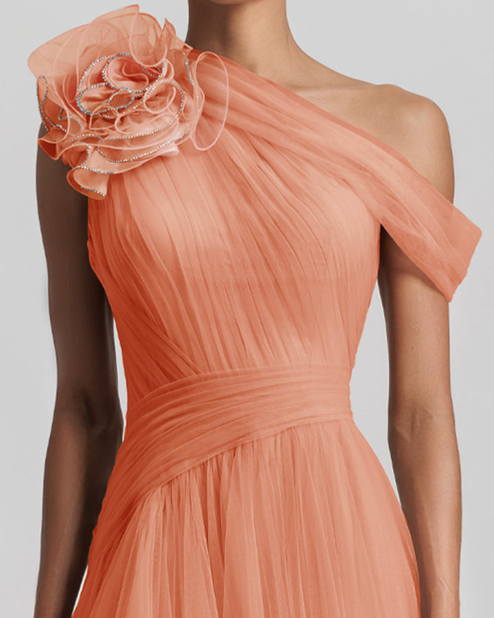 A close-up of an asymmetrical neckline full tulle long evening dress in peach color, with a flower design on the shoulder, and draping design on the bodice.