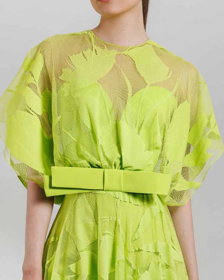 A close-up of a patterned lace midi dress with a bow design on the waist and an asymmetrical hemline, all in lime color.