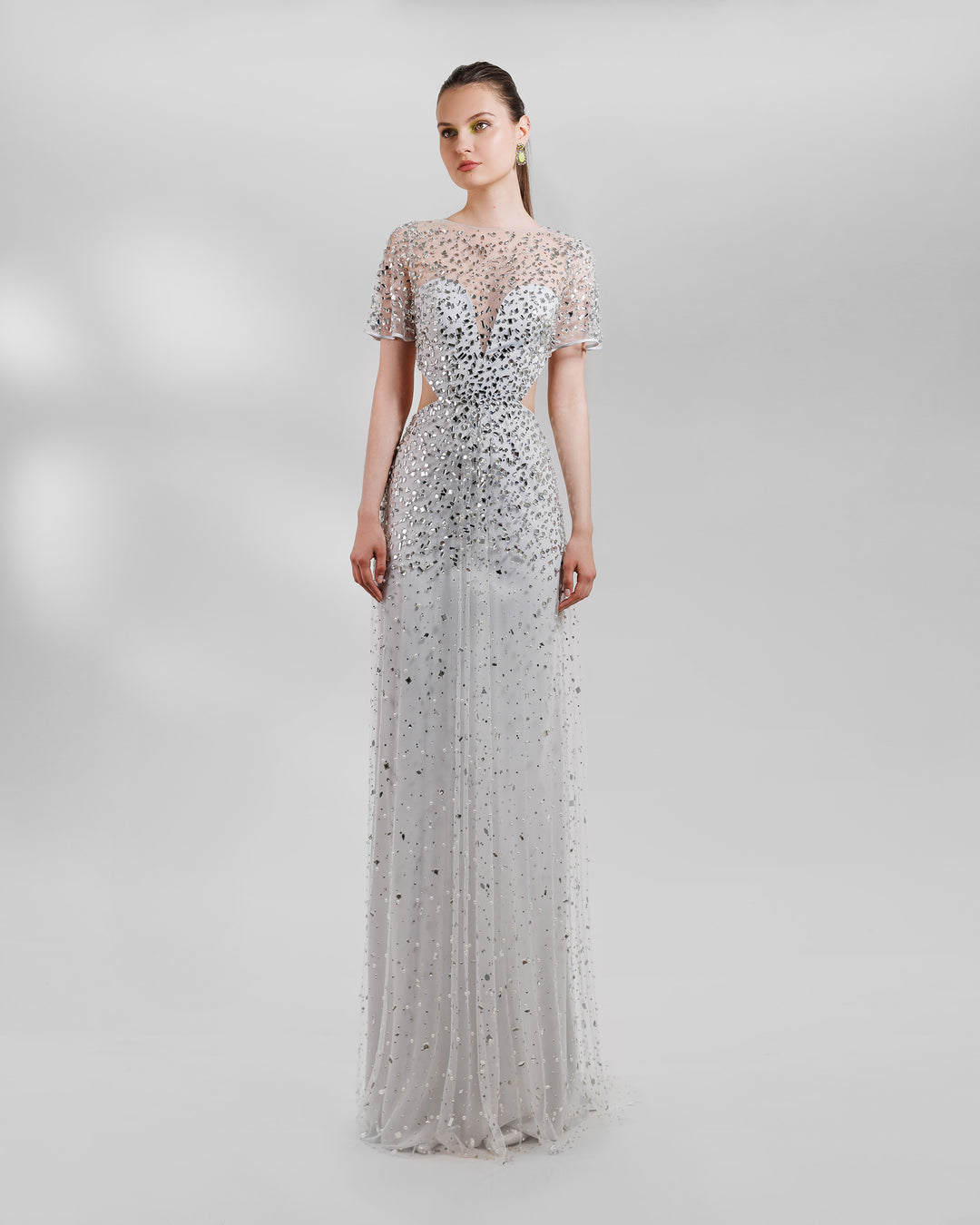 A fully beaded tulle long evening dress, featuring a straight slim cut. Under the tulle a sweetheart shaped bodice with side cut outs, short sheer sleeves and an open back, all in grey color.