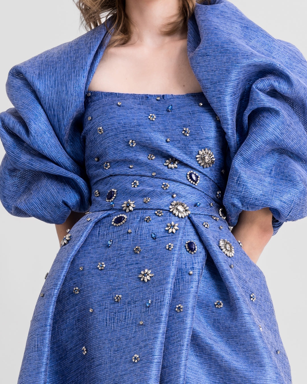 A close-up of a strapless draped blue evening dress with beadings, paired with a draped bolero.