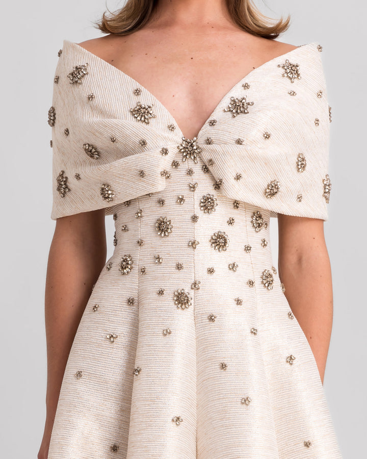 A close-up of an off-the-shoulders cream evening dress with a bow design, beadings, and a draped skirt