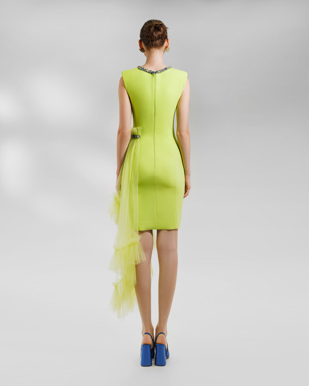 The back of a short structured lime dress with padded shoulders and a round beaded neckline. It features an asymmetrical tulle ruffles on the side with a beaded line.