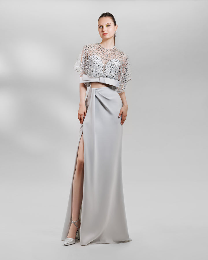 An evening wear grey set featuring a fully beaded top with a crepe bow paired with a draped skirt featuring a slit on the side.