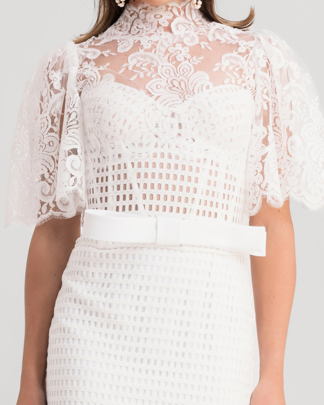 A close-up of a white midi evening dress featuring a high-collar lace top with flared sleeves and a bow design at the waist, paired with a midi lace pencil skirt.