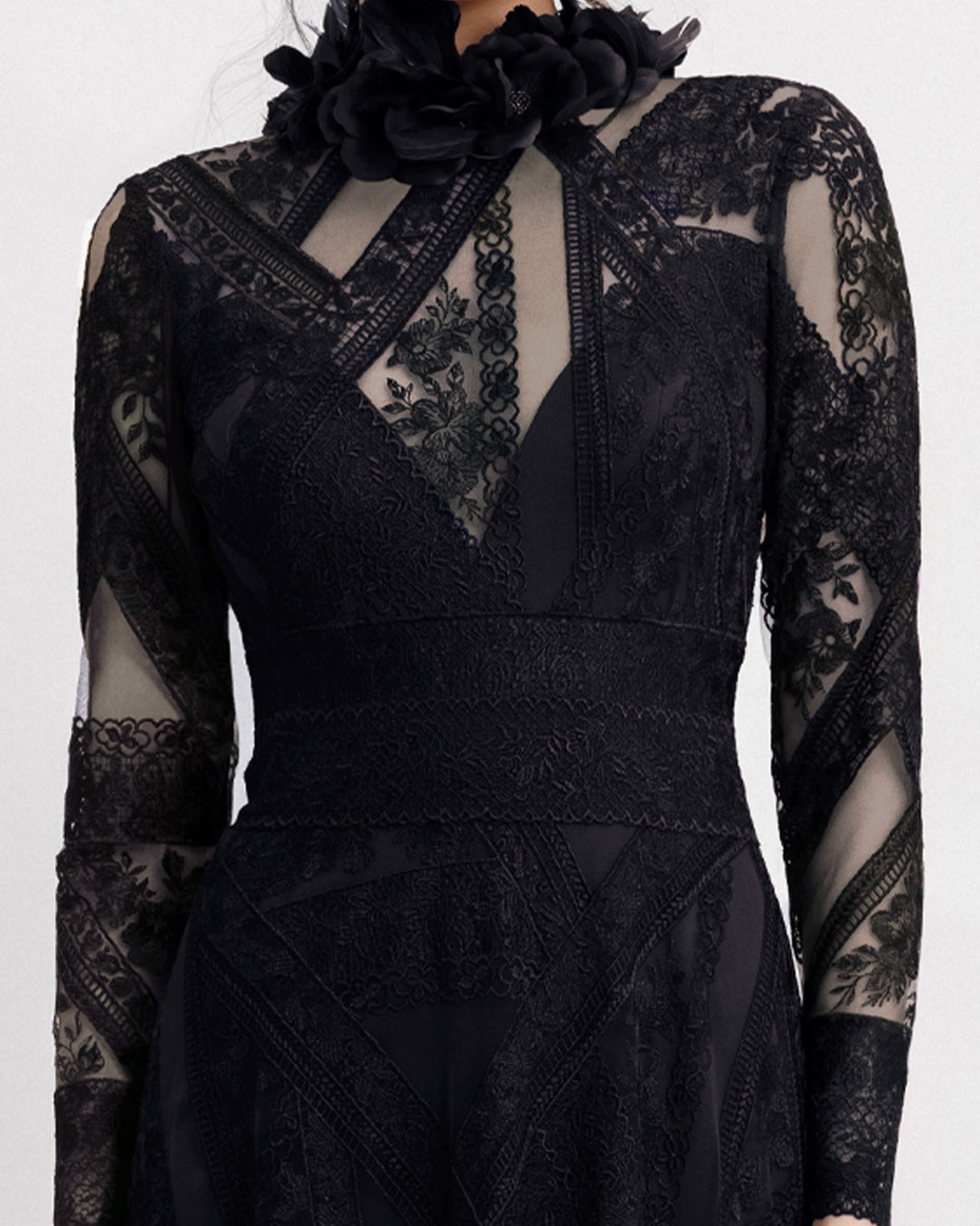 A close-up off  a long sleeved black evening dress in a patterned lace fabric, featuring an embroidered floral feathers on the collar.