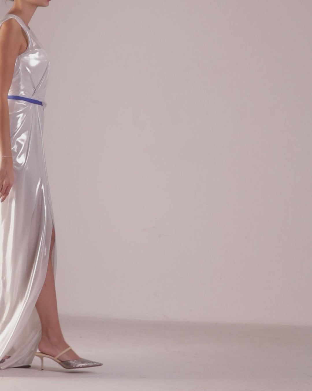 An asymmetrical silver laminated pleated dress with and open back featuring a slit on the side and a contrasting blue belt.