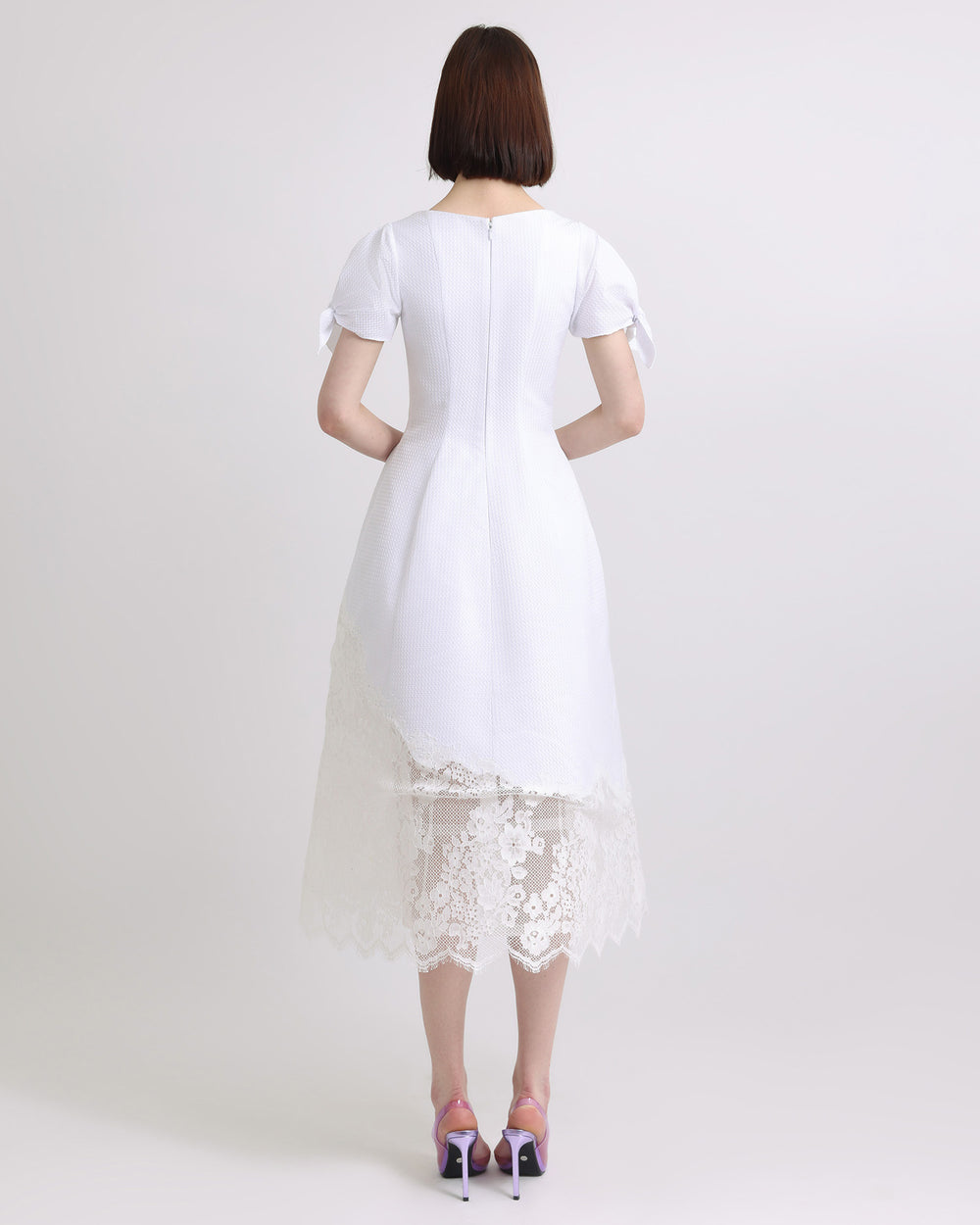 The back of a flared White dress with puffed sleeves and asymmetrical intricate lace on the hemline.