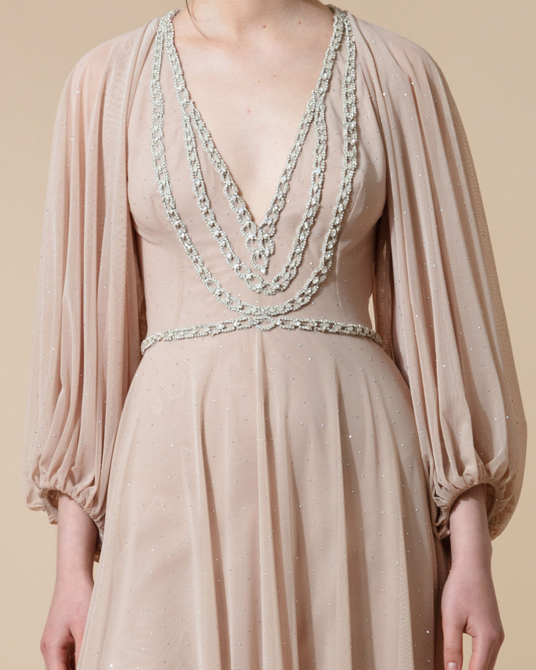 A close-up of a beige evening dress featuring an embellished deep V neckline and long raglan sleeves.