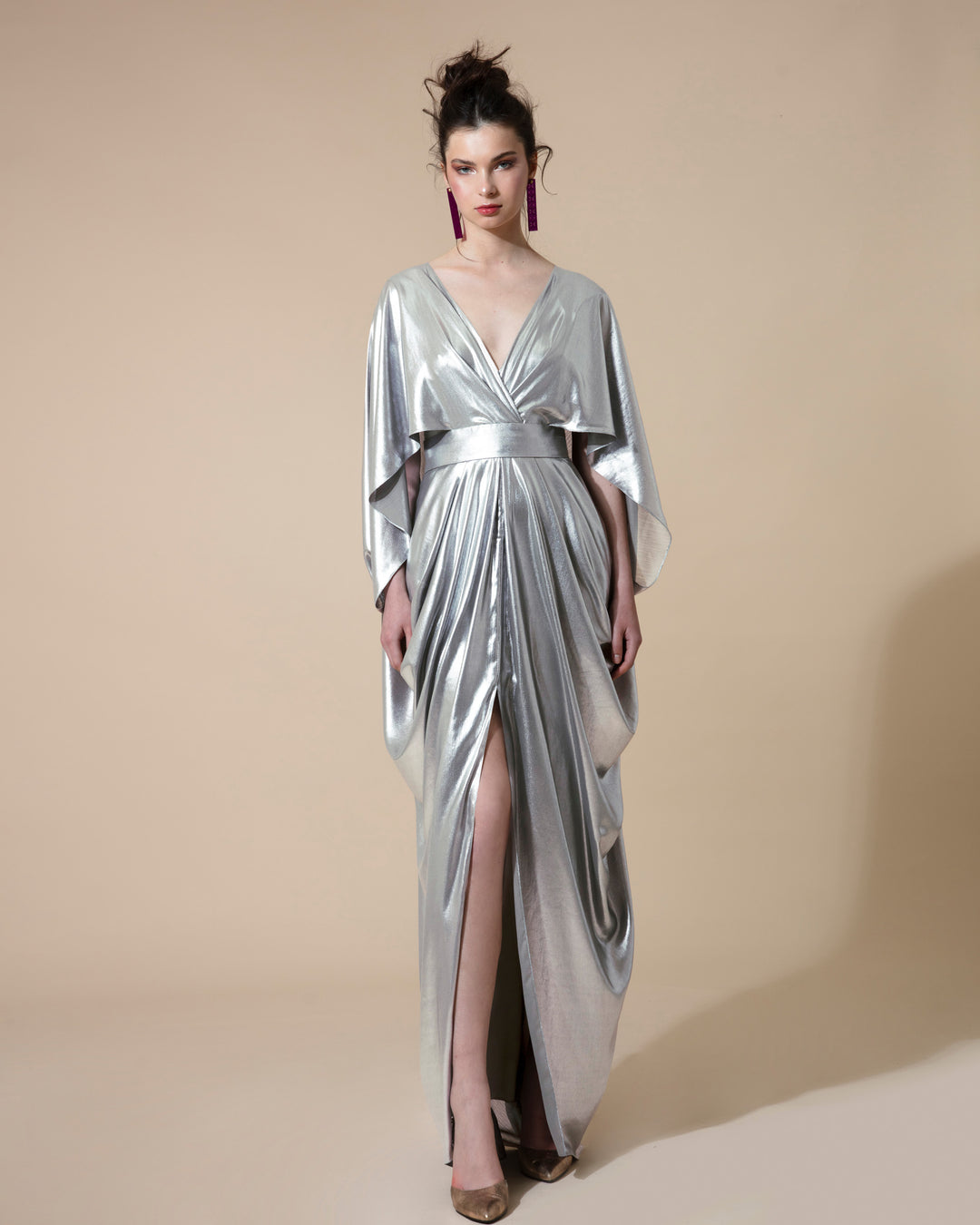 A Cape-like long draped evening dress in metallic silver fabric with a wide V neck, fitted on the waist and an overlapping middle slit.