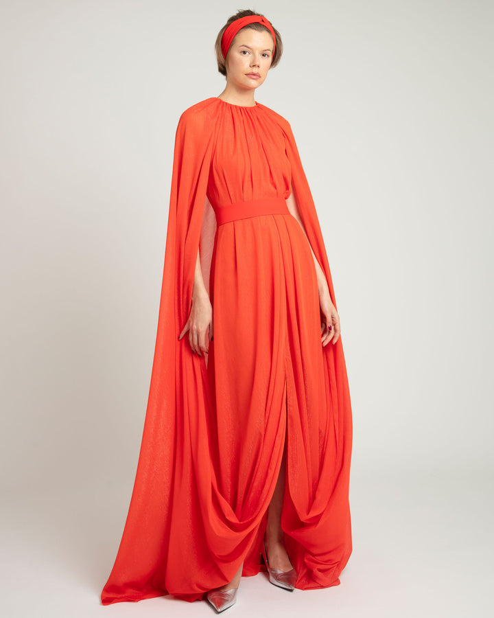 A round neckline kaftan style dress in coral color featuring an overlapped cape-like sleeves, a belt, a middle slit, and draping details.