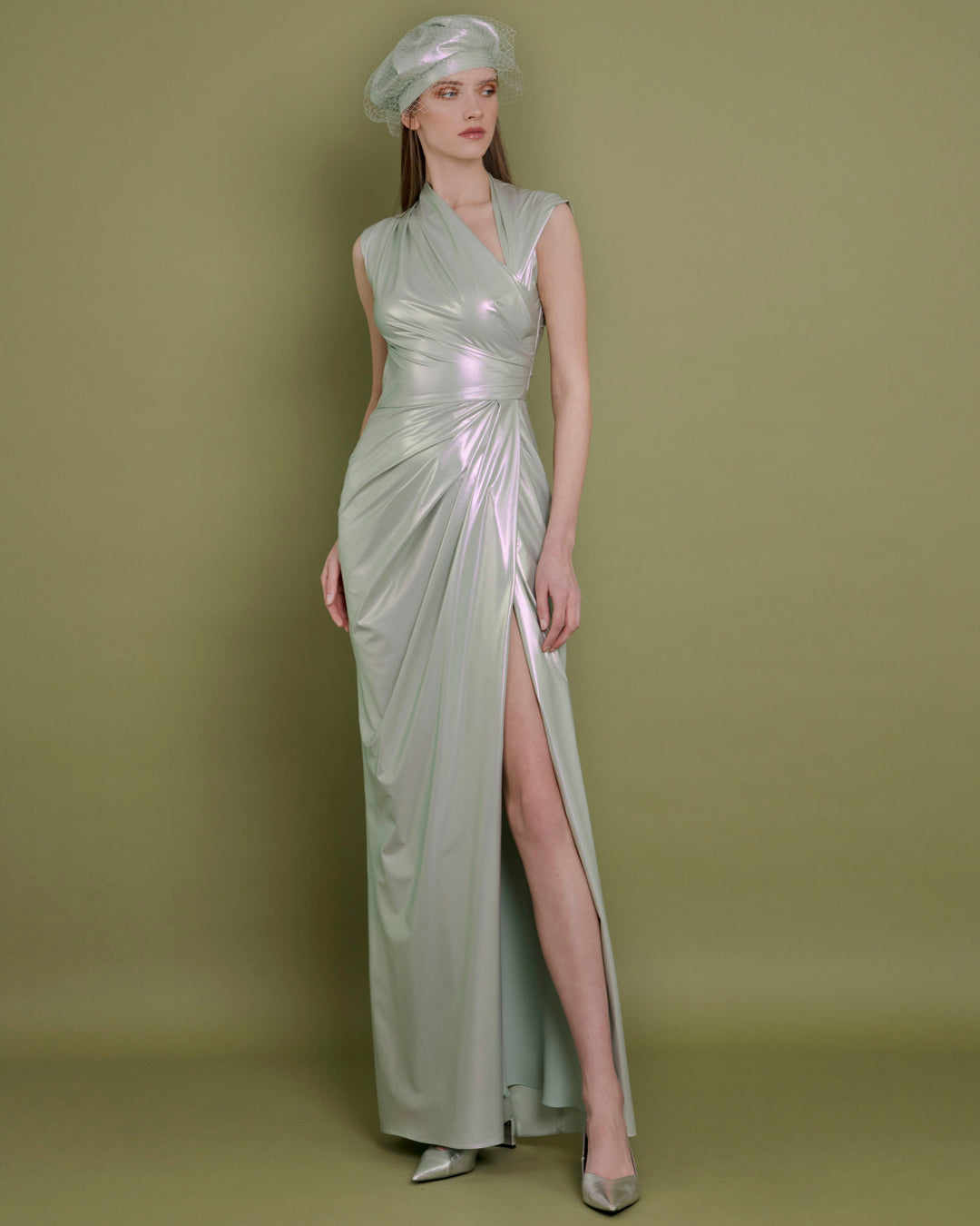 An asymmetrical neckline evening dress in metallic green latex fabric, with draping details on the waist and an open slit on the side.