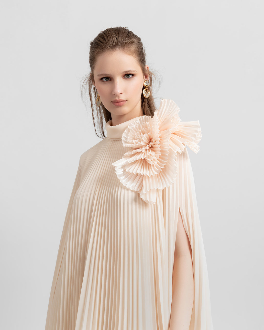 A close-up of a fully pleated lame organza flared beige dress with an embellished draped flower on the shoulder.