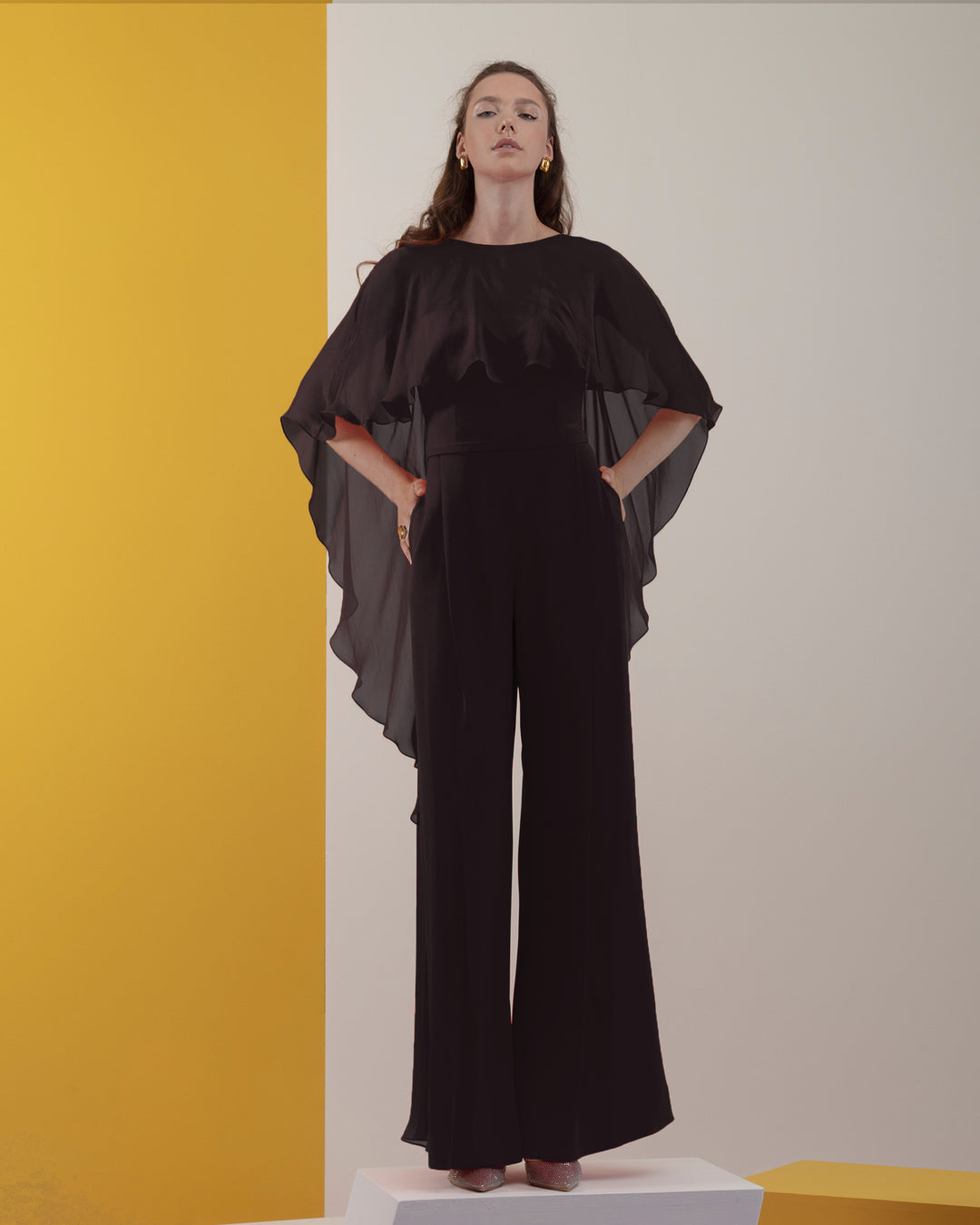 A black jumpsuit with backless design and ruffled flowy chiffon cape-like neckline.