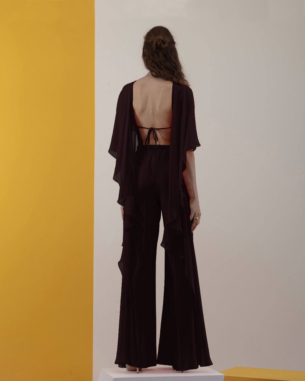 The back of a black jumpsuit with backless design and ruffled flowy chiffon cape-like neckline.