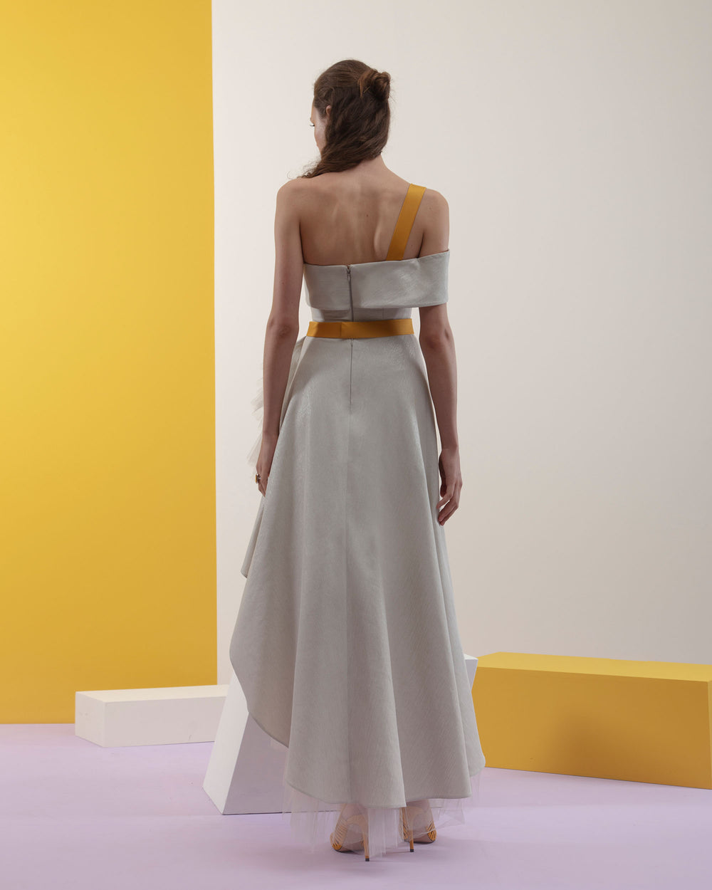 The back of an off-shoulders cut short dress with ruffled tulle underlaying the asymmetrical hemline details in a laminated mikado fabric and a detachable yellow crepe belt and strap.