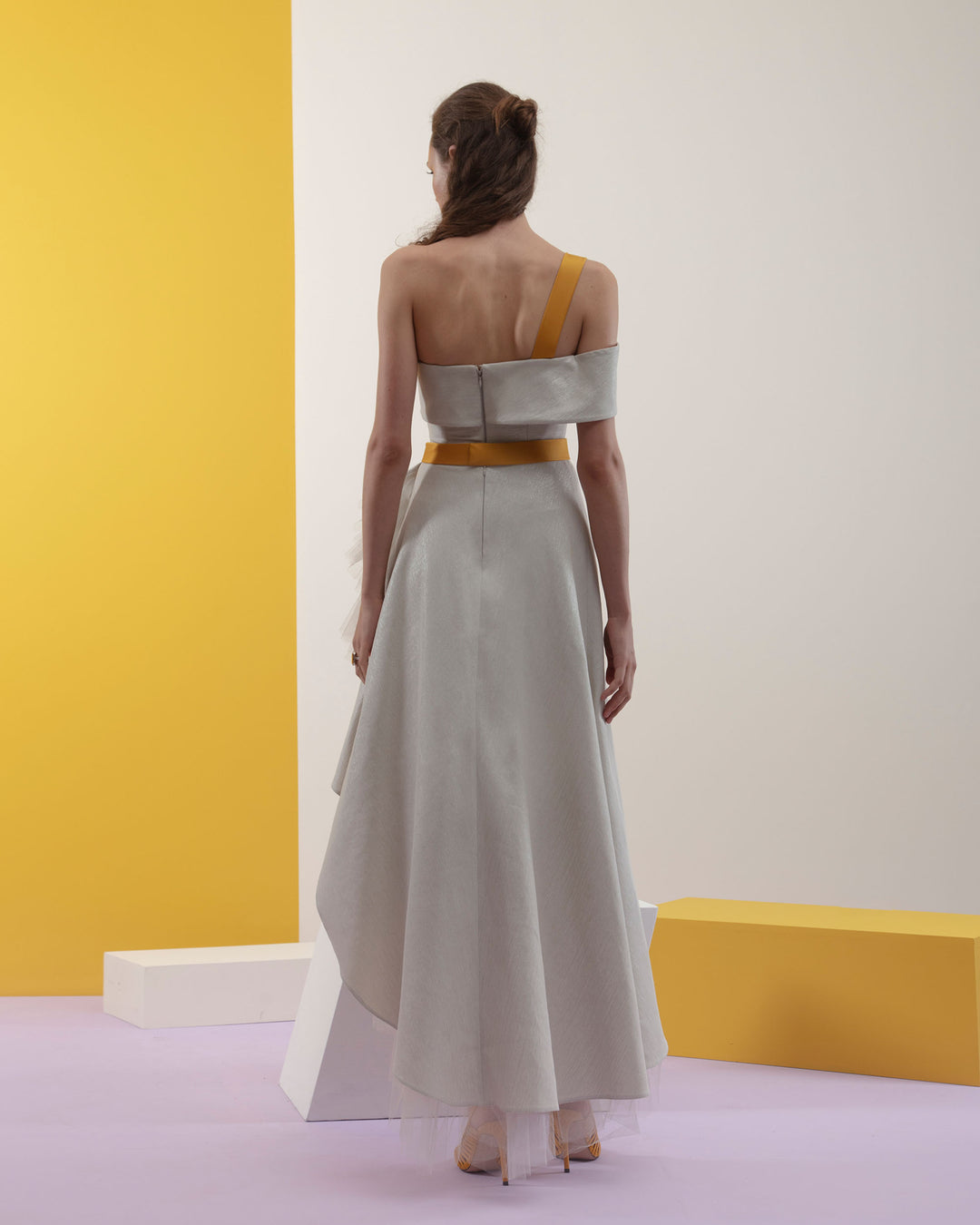 The back of an off-shoulders cut short dress with ruffled tulle underlaying the asymmetrical hemline details in a laminated mikado fabric and a detachable yellow crepe belt and strap.