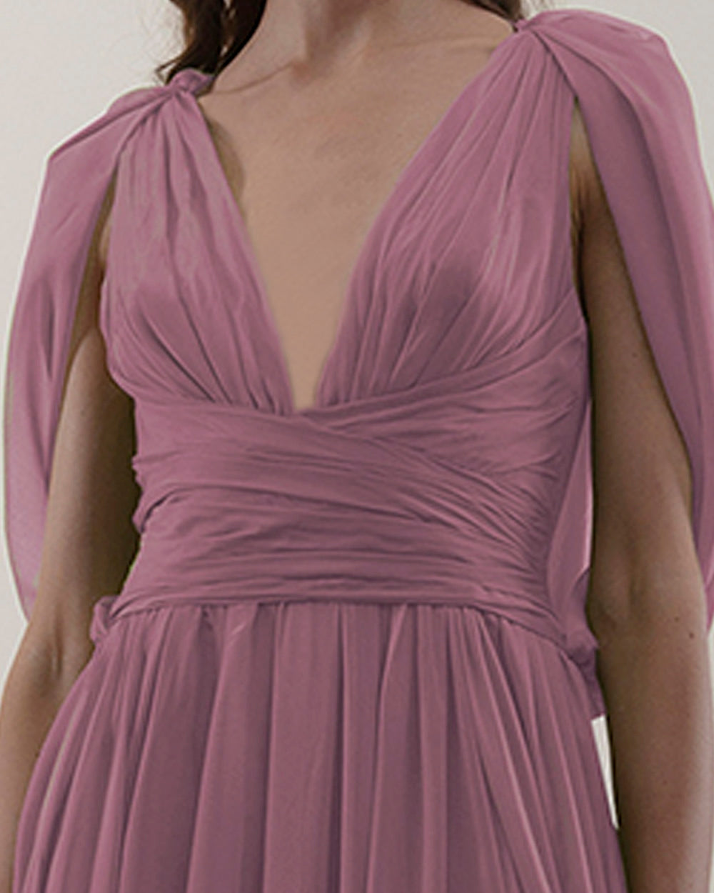 A close-up of a deep V-neckline, pink chiffon fabric long evening dress with cape-like sleeves, draped details on the waist, and an open slit on the side.