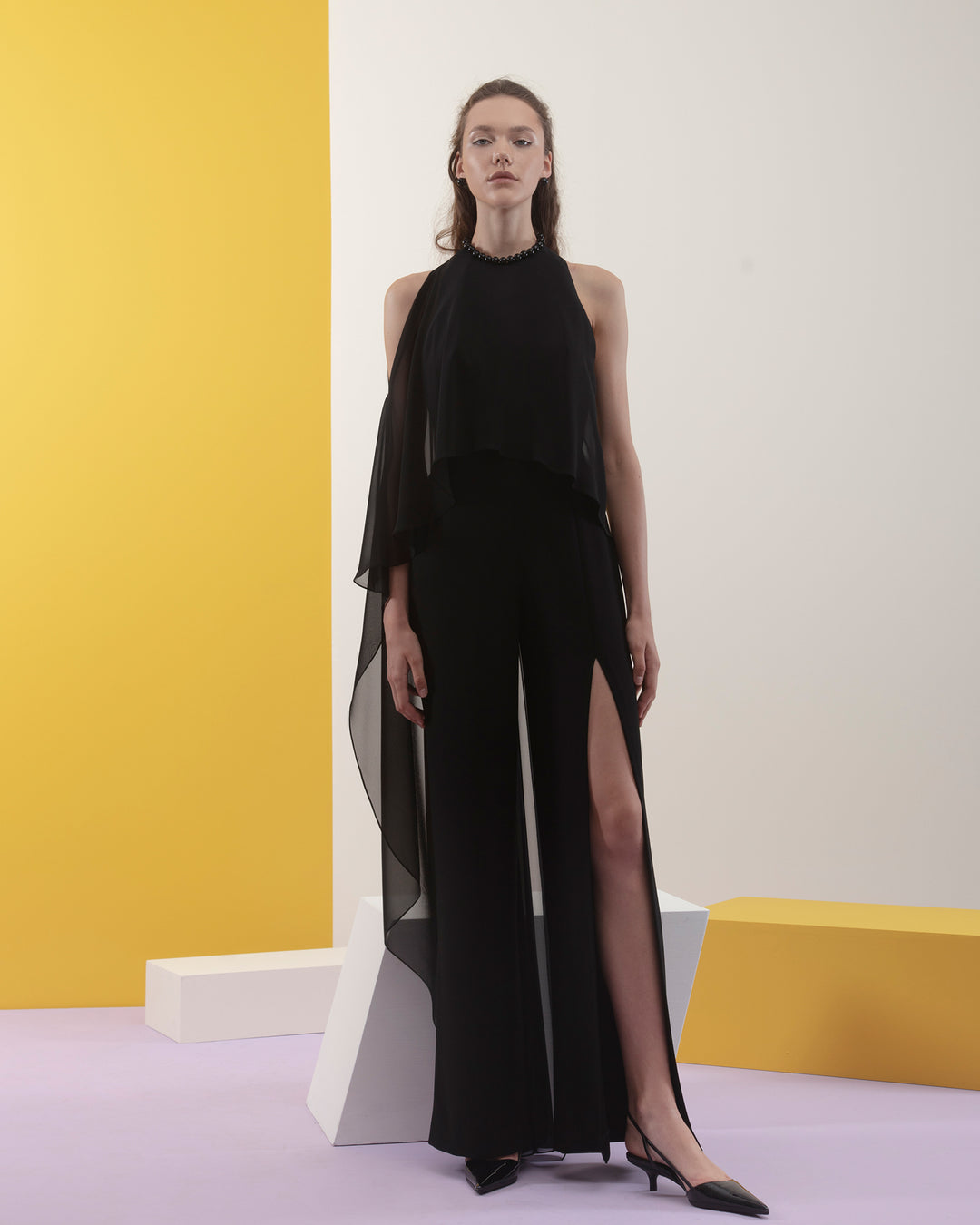 A black crepe jumpsuit with rounded neckline, draped chiffon flowy fabric on the side, and a long slit.