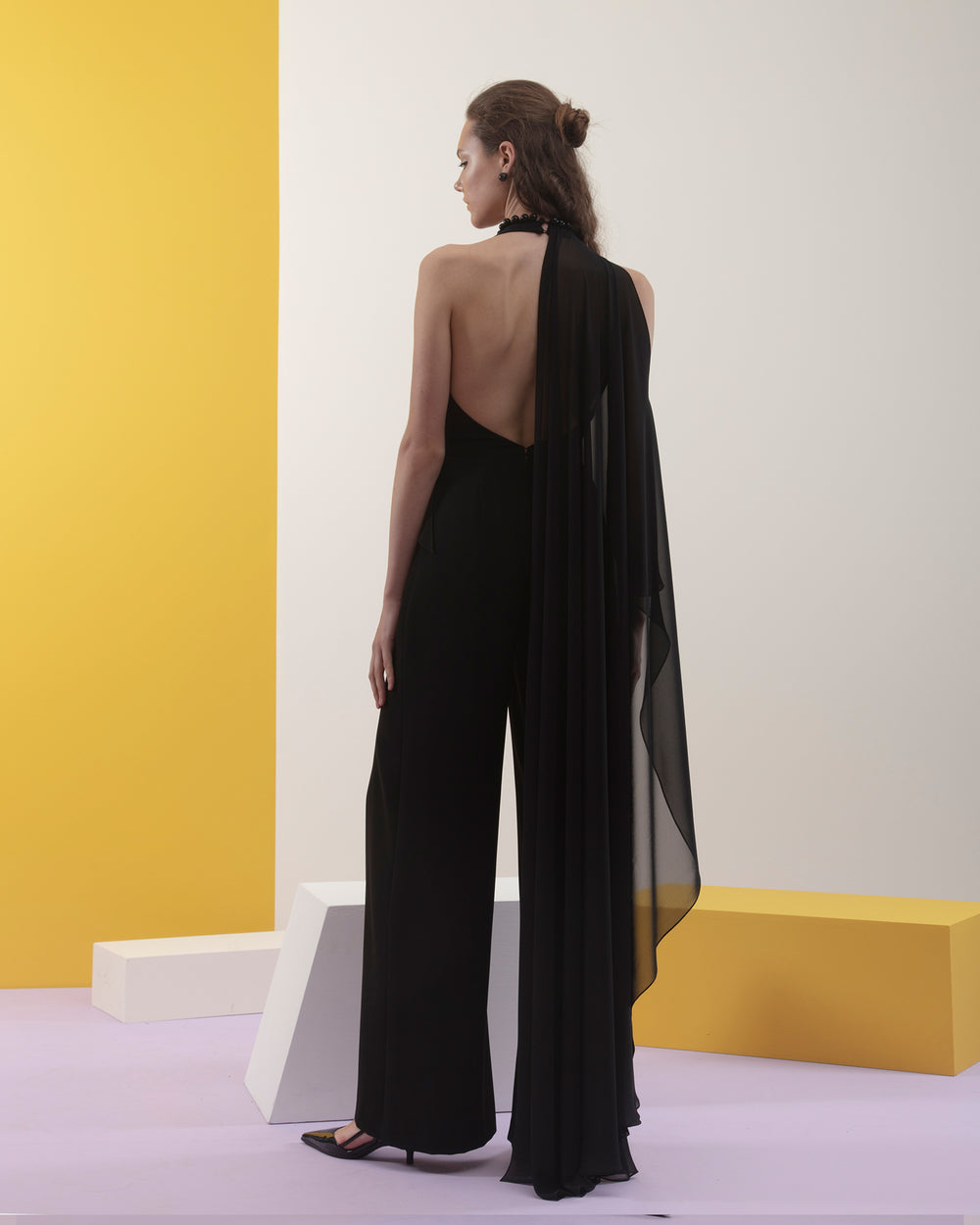 The back of a black jumpsuit with draped shawl like flowy chiffon fabric on the side, and an open back.