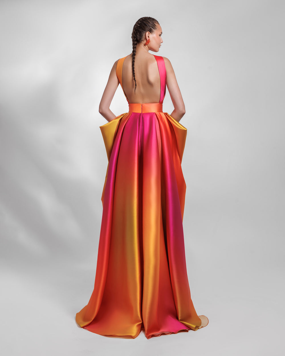 The back of a deep V-neckline heavy satin dress in orange, fuchsia and yellow hues, featuring an open back and a symmetrical pleated overskirt.