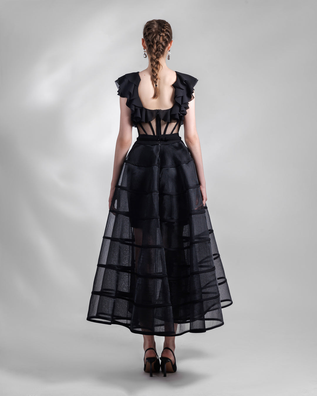 The back of a black evening wear set featuring a ruffled laser cut open back chiffon top with a see-through corseted bodice paired with a cage like long skirt.