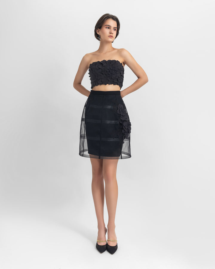 An evening wear set featuring a strapless black cropped-top with laser-cut details, paired with a cage-like black skirt with flower details.