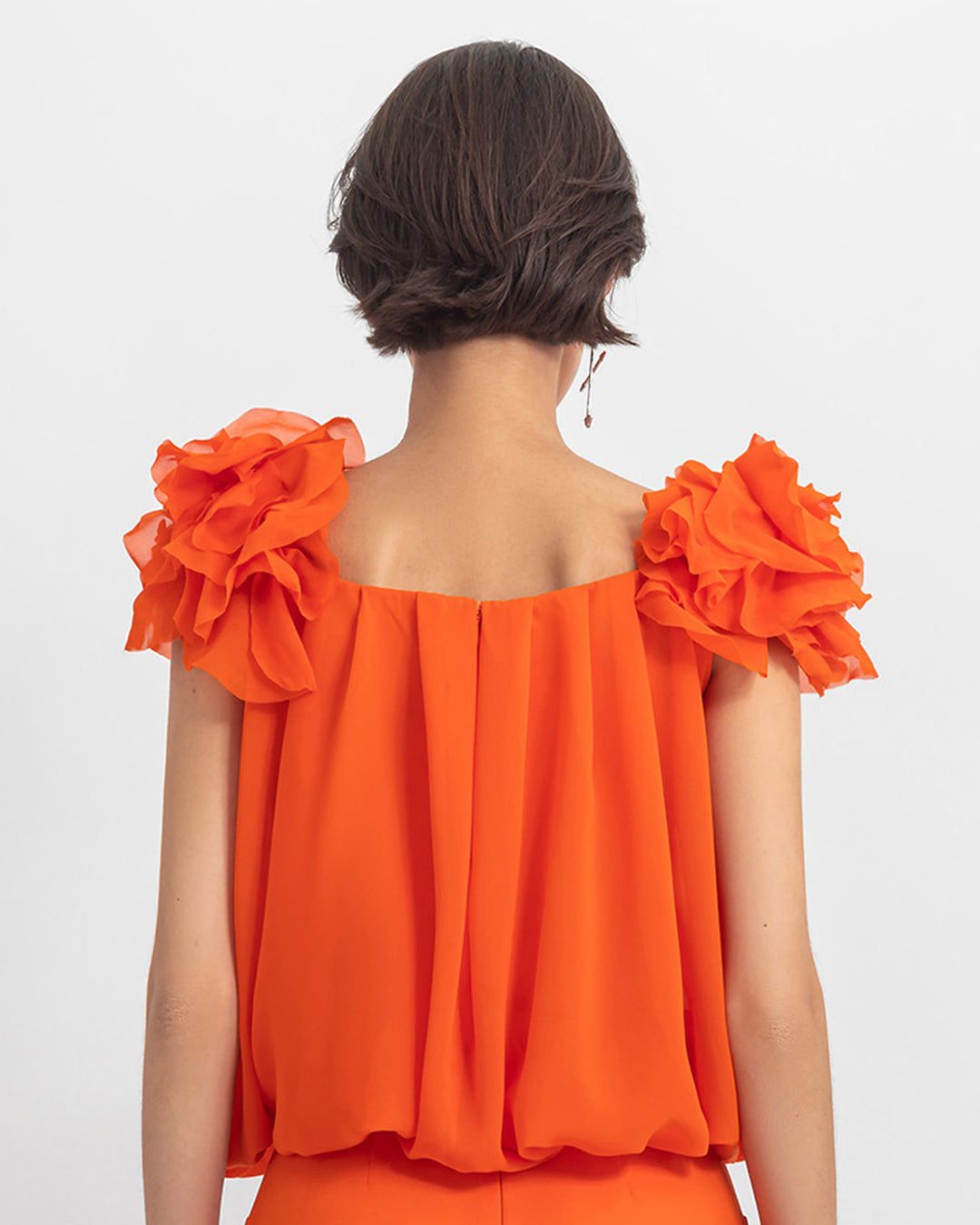 The back of an orange draped loose cut chiffon top with flower details on the shoulders.