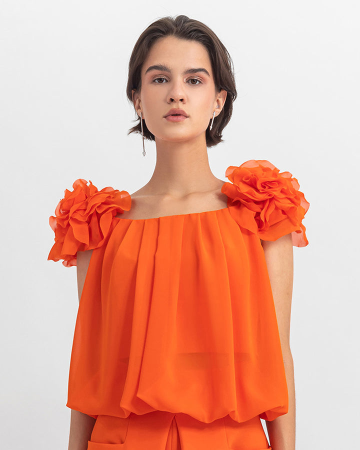 An orange draped loose cut chiffon top with flower details on the shoulders.